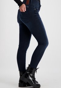 Schlanke Jeans aus dunkel gewaschenem Denim mit hoher Taille, Gesäßtaschen und glatter Textur. Kombiniert mit glänzenden, schwarzen Schnürstiefeln.