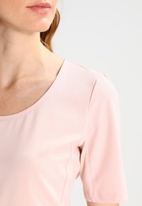 Top rosa claro de manga corta con cuello redondeado, textura suave y detalles sutiles en los hombros, que muestra una silueta ajustada.