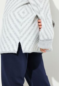 Pull en tricot rayé gris et blanc, avec une coupe décontractée, des fentes sur les côtés et des poignets côtelés, associé à un pantalon bleu marine. Texture douce visible.