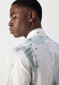 Ανθεκτικό πουκάμισο με κουμπιά σε λευκό και παστέλ πράσινο, διαθέτοντας υφασμάτινη υφή και όρθιο γιακά. Η πίσω όψη δείχνει λεπτομέρειες στους ώμους.