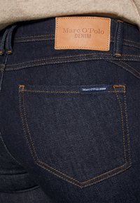 Jeans en denim bleu foncé avec une étiquette en cuir marron portant la mention "Marc O'Polo DENIM" et des détails de couture orange sur les poches arrière.