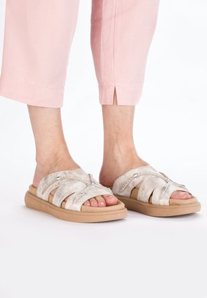 Light beige sandals with crisscross suede straps, featuring decorative rhinestones, and a cushioned, tan rubber sole.