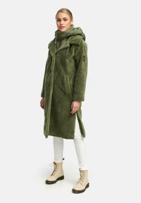 Lang groene fleecejas met capuchon, voorzakken en zijsplitten. Gepaard met crèmekleurige veterschoenen en witte broek.