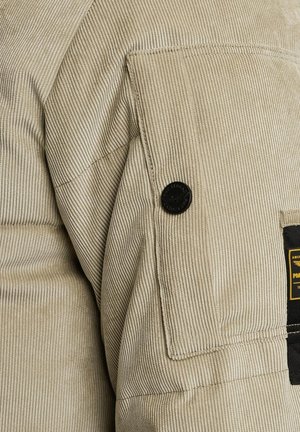 Tissu en velours côtelé beige avec des rayures verticales, comportant un bouton noir et une poche plaquée avec une étiquette sur le côté. Couture détaillée visible.