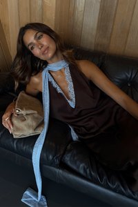 Abito di raso marrone con scollatura in pizzo, sciarpa in pizzo blu chiaro annodata al collo, mentre tiene una borsa in suede beige, seduta su un divano in pelle nera.