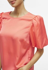 Blusa di raso corallo con scollatura rotonda e maniche a sbuffo. Texture liscia con una leggera lucentezza e dettaglio della tasca laterale.