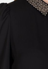 Blusa nera con colletto texturizzato caratterizzato da dettagli in perline. Le maniche a palloncino e il tessuto morbido creano una silhouette elegante.
