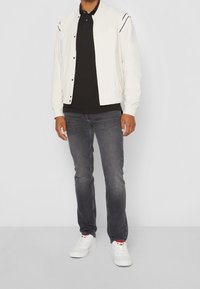 Veste bomber créme avec des boutons, polo noir, jeans gris et baskets blanches avec des accents rouges. Tenue décontractée avec des couleurs contrastantes.