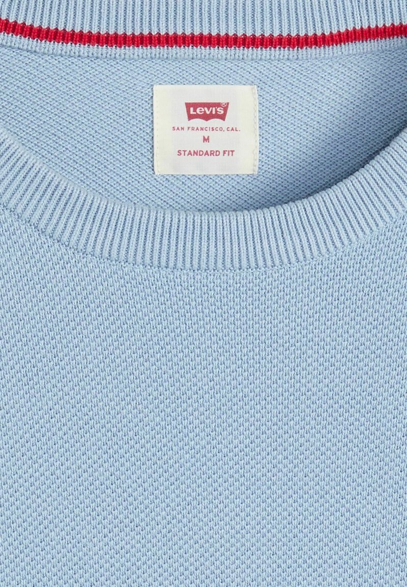 Cuello de suéter tejido en azul claro con cuello acanalado, detalle de franjas rojas y blancas, y etiqueta Levi's Standard Fit talla M en el interior.