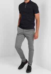 Marinblå polka-prickig pikétröja med ficka, matchad med ljusgrå slim-fit byxor och svarta sneakers, på en neutral bakgrund.