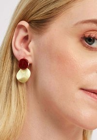 Rdeče in zlato uhane z bleščečo rdečo krožno zgornjo obliko in brušenim zlatenim ovalom spodaj, kar ustvarja kontrast v teksturi in barvi.