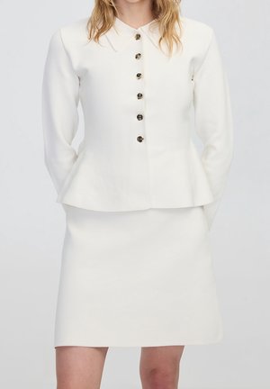 Femme portant une veste blanche à manches longues avec un col pointu, sept boutons, et une jupe assortie jusqu'aux genoux sur un fond clair.