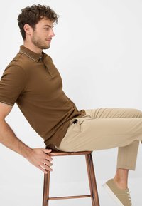 Polo marrone con una patta a due bottoni e colletto a contrasto, abbinato a pantaloni beige chiaro. Il modello è seduto su uno sgabello da bar in rame.