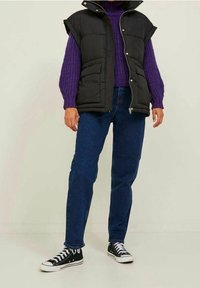 Černá péřová vesta s vysokým límcem, fialový pletený svetr, modré džíny a černé tenisky s vysokým kotníkem. Pohodlný vrstvený outfit.