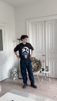 T-shirt gráfica preta com o rosto de uma pessoa, combinado com calças azuis de cordão. O cenário inclui paredes brancas e uma mesa com padrão.