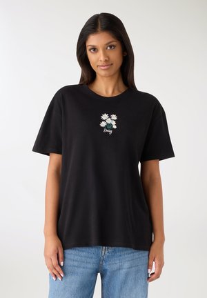 Giovane donna che indossa una t-shirt nera con fiori di margherita bianchi e la scritta "Daisy", abbinata a jeans azzurri, in piedi davanti a uno sfondo bianco.