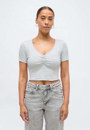 Barna haját kontyba fogó nő ezüst karika fülbevalót visel, világosszürke, ráncolt rövid ujjú crop topot és magas derekú, kifakult szürke farmert visel.