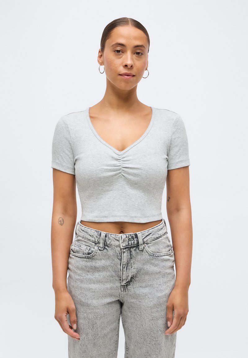 Γυναίκα με καστανά μαλλιά σε κότσο φοράει ασημένια κρίκους σκουλαρίκια, ανοιχτό γκρι κοντομάνικη ρούά μπλούζα με πιέτες και ψηλόμεσα ξεθωριασμένα γκρι τζιν.