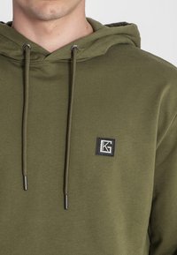 Felpa verde oliva con design a cordoncino, caratterizzata da una patch quadrata nera con il logo "KG" sul petto e dettagli metallici argentati.