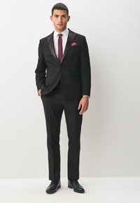Schwarzer Anzug mit satinierter Rever, weißem Hemd, kastanienbraunem Schlips und Einstecktuch. Schmal geschnittene Hose und schwarze Lederschuhe. Schlichter Hintergrund.