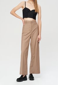 Donna che indossa un crop top bustier nero, pantaloni beige a vita alta e larghi, e mocassini neri spessi, in piedi con una mano sui fianchi.