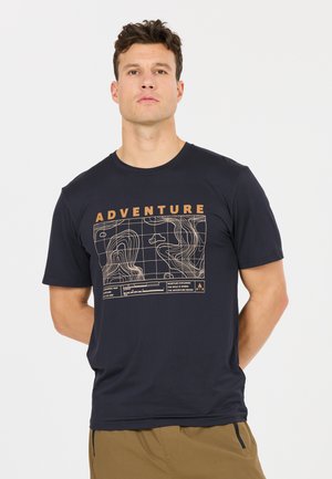 Mann trägt ein schwarzes T-Shirt mit topografischer Karte und dem Schriftzug "ADVENTURE", kombiniert mit braunen Hosen, steht vor weißem Hintergrund.