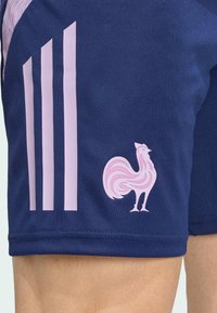 Pantalones cortos deportivos de color azul marino con acentos en rosa, que presentan tres rayas verticales y un logotipo de gallo estilizado en la pierna izquierda. Textura suave.