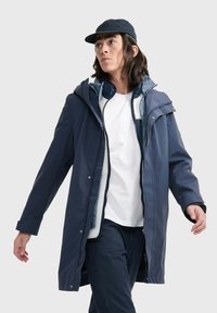 Veste imperméable marine avec capuche, fermeture éclair à l'avant et accents à bouton-pression. Doublée d'un motif texturé ; associée à une chemise blanche.