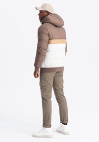 Chaqueta tipo puffer en marrón, crema y beige con capucha, dobladillo acanalado y costuras grandes. Combinada con pantalones cargo y zapatillas blancas.