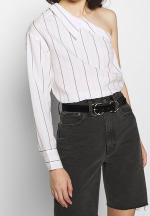 Asymmetrisk hvid bluse med sorte lodrette striber og enkelt skulderdesign. Kombinationen består af shorts i sort denim med høj talje og et matchende bælte.