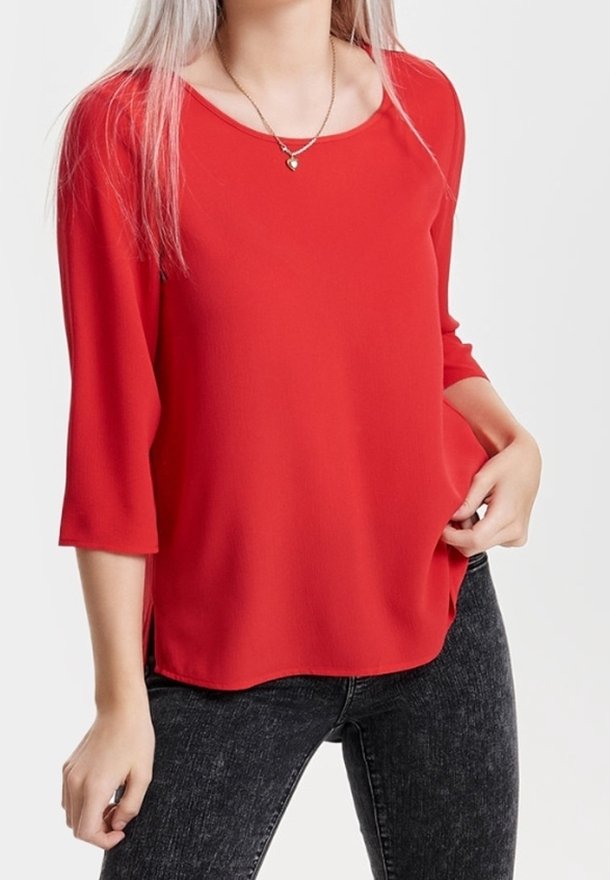 Femme portant un chemisier ample rouge vif à manches trois-quarts et un jean noir délavé, accessoirisée d'un petit collier pendentif.
