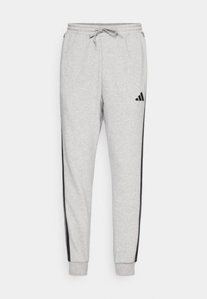 Pantalones deportivos Adidas gris claro con franjas laterales negras, una cintura elástica con cordón y un logo negro de Adidas en el muslo izquierdo.