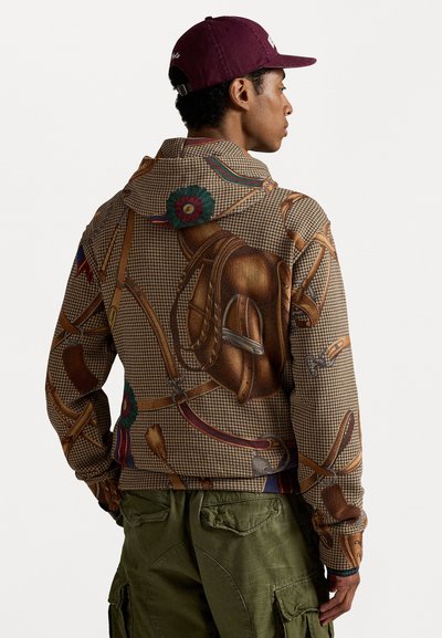 Homem de hoodie castanho com padrão de arreios de cavalo, calças cargo verdes-oliva e um boné de basebol cor bordô, virado de lado.