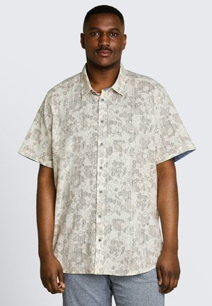 Man staat, draagt een beige overhemd met korte mouwen met abstract patroon en grijze broek, neutrale achtergrond.