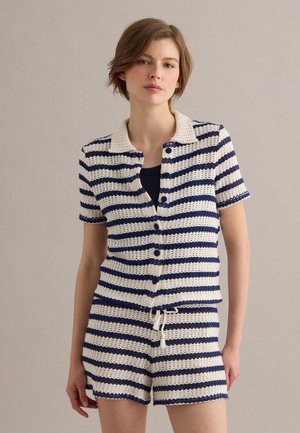 Joven mujer con camiseta de punto de manga corta con botones y rayas horizontales en azul marino y blanco, y pantalones cortos a juego, de pie frente a un fondo liso.