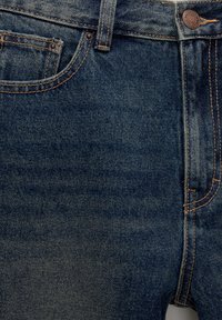 Gros plan sur le devant d'un jean en denim bleu avec poche visible, passant de ceinture, bouton et détails de couture orange.