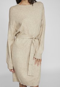 Beige stickad klänning med lång ärm, bälte i midjan, ribbade mudd och en avslappnad passform. Mjuk textur med en mysig känsla.