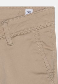 Zbliżenie na beżowe spodnie, ukazujące szlufki na pasek, przednią kieszeń oraz widoczny metkę rozmiaru "128" wewnątrz pasa.