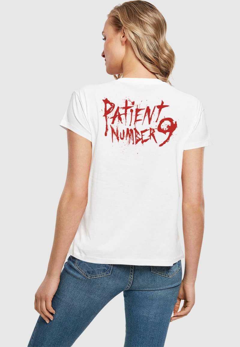 T-shirt blanc à manches courtes avec un texte graphique rouge "NUMÉRO DE PATIENT 9" au dos, avec un effet de éclaboussures. Design ajusté.
