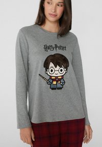 Camisa gris de manga larga con un diseño caricaturesco de Harry Potter. Texto en la parte superior que dice "Harry Potter". Llevada con pantalones de pijama de cuadros rojos.