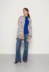 Lang beige skjorte med sort bølgemønster, båret over en blå top. Udflydende blå jeans og sorte hælede sandaler fuldender outfittet.