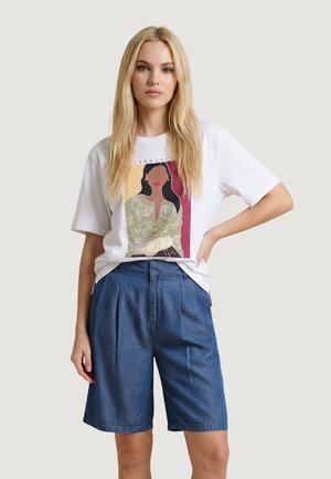 Donna bionda che indossa una t-shirt grafica bianca con stampa astratta femminile e pantaloncini blu a vita alta e lunghezza al ginocchio, in piedi con una mano sui fianchi.