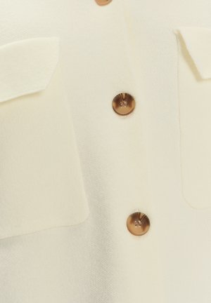 Camisa de textura cremosa con dos bolsillos frontales y botones marrones. La tela parece suave, con un color sutil y uniforme.