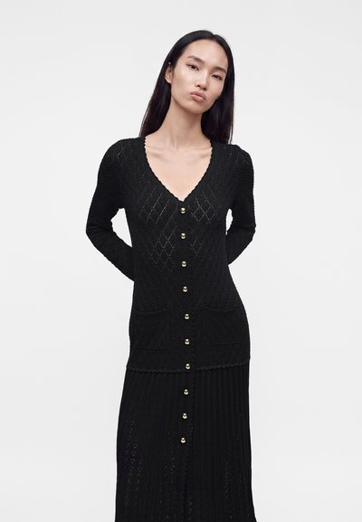 Femme aux longs cheveux foncés portant une robe cardigan en maille noire texturée avec des boutons dorés, debout devant un fond blanc uni.