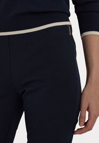 Mörknavy byxor har en elegant design med en ribbad midja och en kontrasterande ljusfärgad kant. En liten gulddragkedja är synlig som en accent.