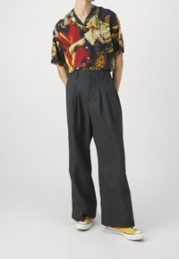 Camisa de botón de mangas cortas con un estampado artístico multicolor; combinada con pantalones oscuros de pierna ancha y cintura alta, y zapatillas amarillas.
