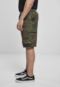 Pantalones cortos cargo en un patrón de camuflaje verde, hechos de tela ligera con bolsillos laterales, combinados con zapatillas negras y blancas.