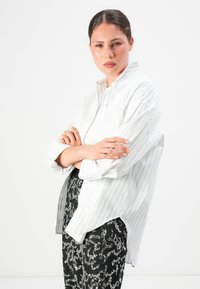 Sieviete ar rokām sarautām, vilkusi baltu svītrainu krekli un melnus mirdzošus bikses, stāv pretī vienkāršai baltai fona.