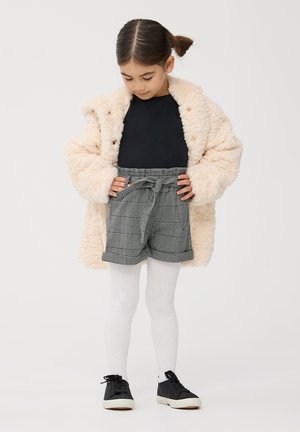 Cappotto beige morbido, top nero, pantaloni corti grigi a quadri con vita regolabile, collant bianchi e sneakers nere. La modella guarda in basso verso l'outfit.