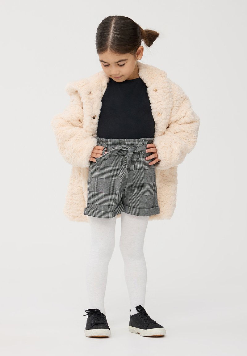 Cappotto beige morbido, top nero, pantaloni corti grigi a quadri con vita regolabile, collant bianchi e sneakers nere. La modella guarda in basso verso l'outfit.